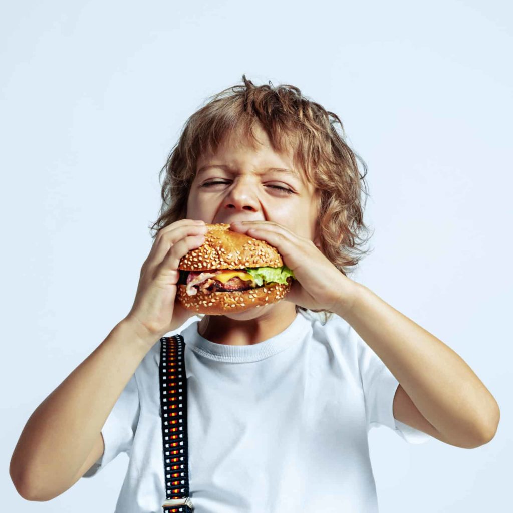Young school boy eating hamburger on light blue background